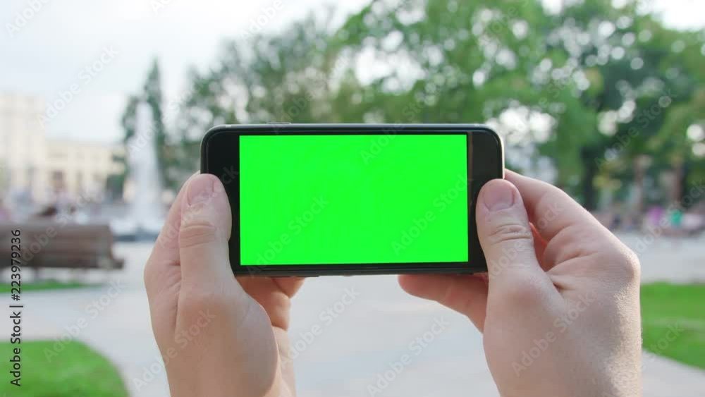 Two hands holding a phone with a green screen in a horizontal position in a public park. Close-up shot. Soft focus Two hands holding a phone with a green screen in a horizontal position in a public park. Close-up shot. Soft focus