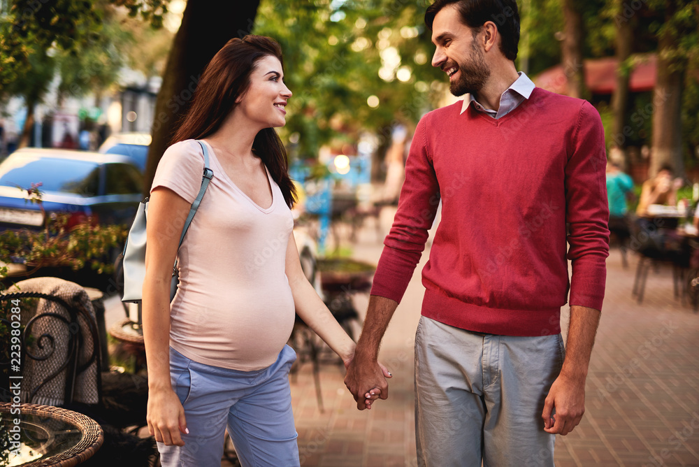 Spending time together. Portrait of happy bearded man holding hand of his beautiful pregnant wife. Sidewalk, cars and trees on blurred background