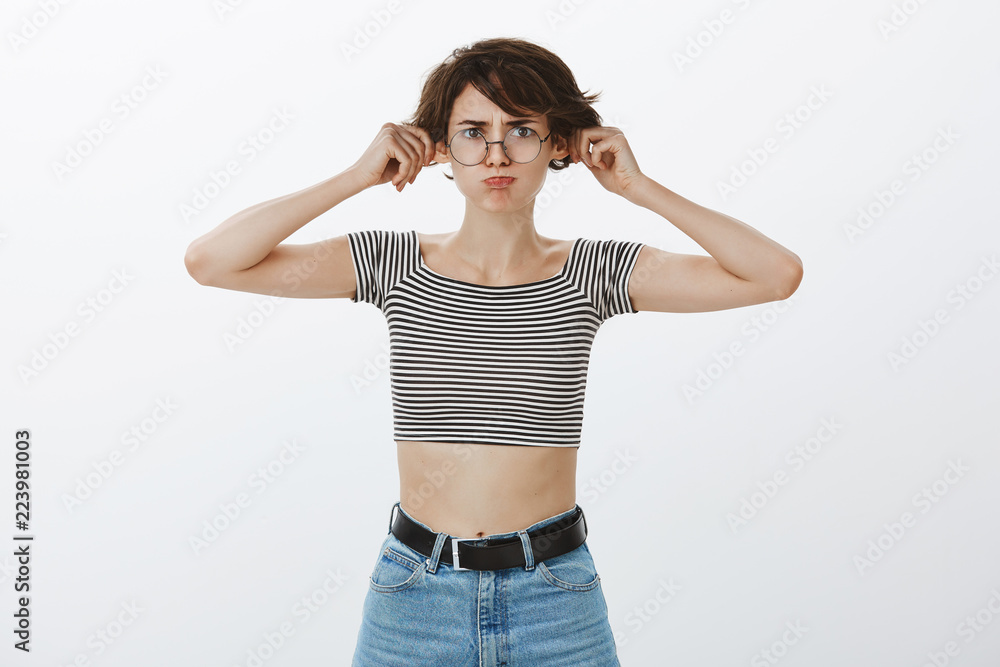 Portrait of funny emotive young european woman in trendy round glasses and striped cropped top, pulling ears with raised hands, pouting and frowning, being childish and careless Portrait of funny emotive young european woman in trendy round glasses and striped cropped top, pulling ears with raised hands, pouting and frowning, being childish and careless