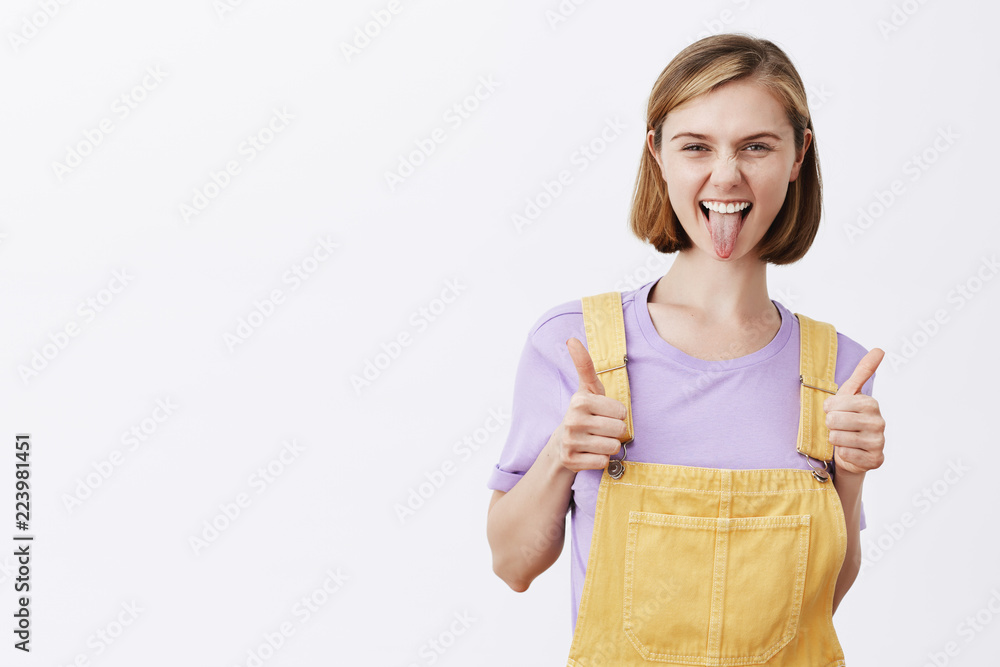I like my new outfit. Pleased excited attractive female with blond hair, holding hands on dungarees and showing thumbs up, sticking out tongue joufully and frowning while liking idea and saying yes I like my new outfit. Pleased excited attractive female with blond hair, holding hands on dungarees and showing thumbs up, sticking out tongue joufully and frowning while liking idea and saying yes