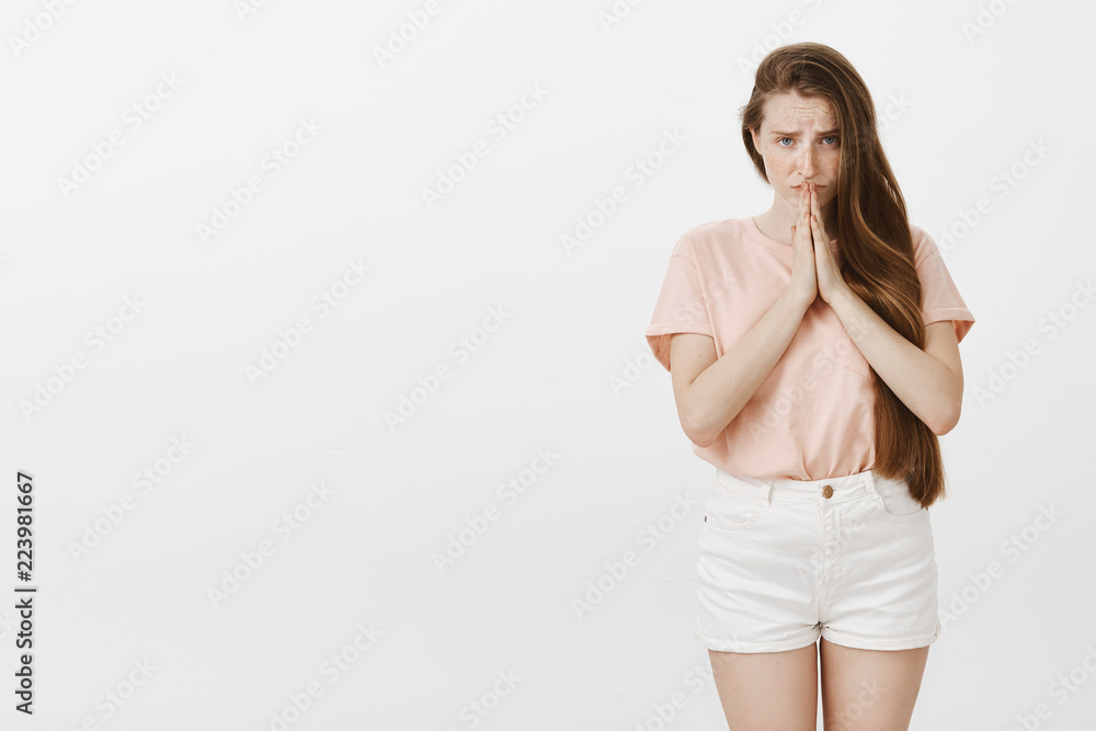 Good-looking desperate and worried caucasian female with long pretty hair, looking from under forehead with silly begging expression, holding hands in pray, asking apology or favor over grey wall