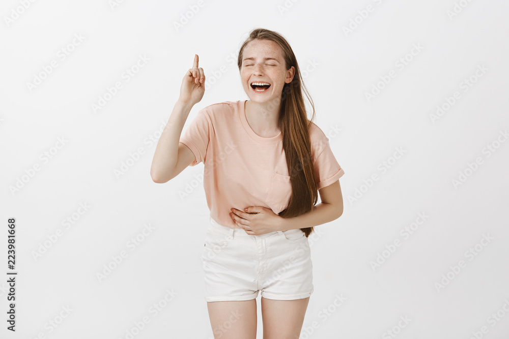 Pretty carefree girl having fun, laughing out loud seeing amusing comedy show, bending from laugh and touching belly, smiling broadly with closed eyes, pointing up with index finger over gray wall Pretty carefree girl having fun, laughing out loud seeing amusing comedy show, bending from laugh and touching belly, smiling broadly with closed eyes, pointing up with index finger over gray wall