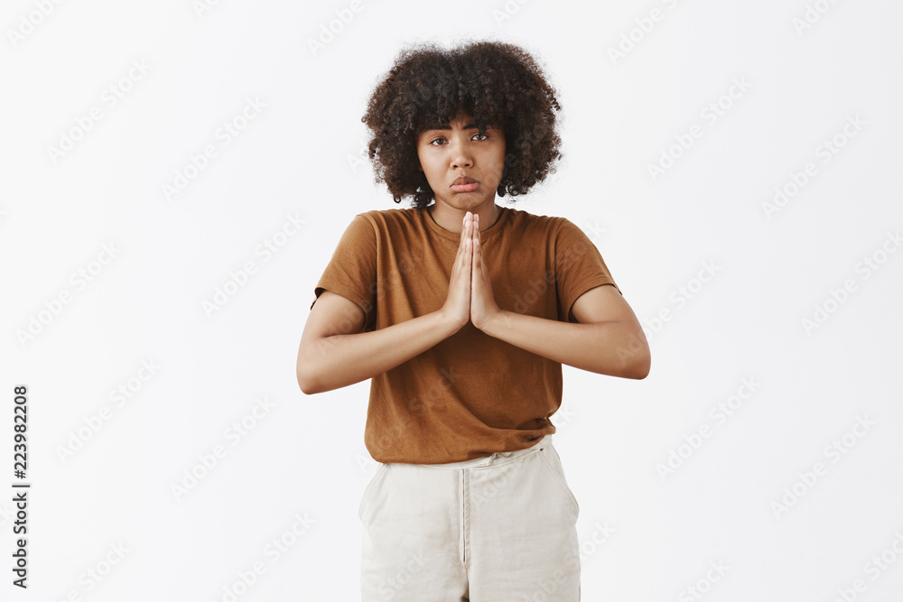 Studio shot of gloomy upset good-looking dark-skinned teenage woman with afro hairstyle pursing lips in sad smile frowning and holding hands in pray while asking help or favor over gray wall Studio shot of gloomy upset good-looking dark-skinned teenage woman with afro hairstyle pursing lips in sad smile frowning and holding hands in pray while asking help or favor over gray wall