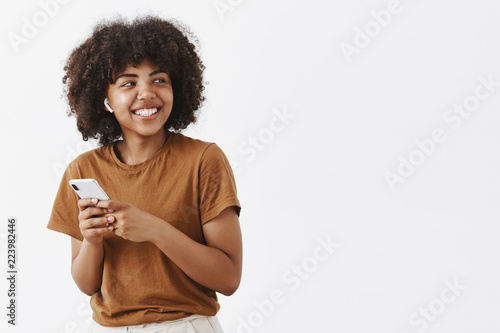 Studio shot of carefree friendly and creative stylish african american teenage girl in brown t-shirt turning right with broad satisfied smile wearing wireless earphones and holding smartphone