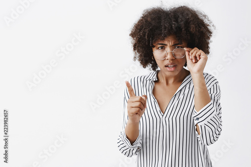 Indoor shot of intense confused and displeased female dark-skinned teacher with afro hairstyle looking from under glasses touching rim and shaking index finger in scolding gesture being disapponted photo