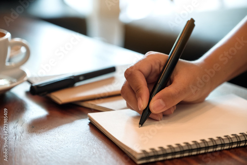 Closeup image of a hand writing down on a white blank notebook with coffee cup on wooden table