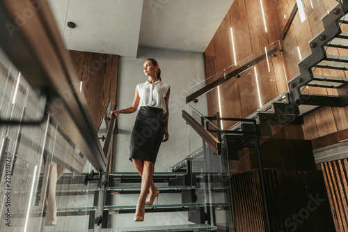 Full length side view orderly elegant businesswoman moving on stairs in wide corridor while looking away inside