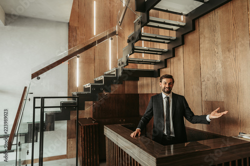 Portrait of happy unshaven male employee gesticulating hand while locating at counter in hotel