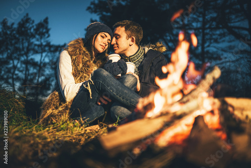 Pretty young couple drinking hot beverage in the forest near bonfire.