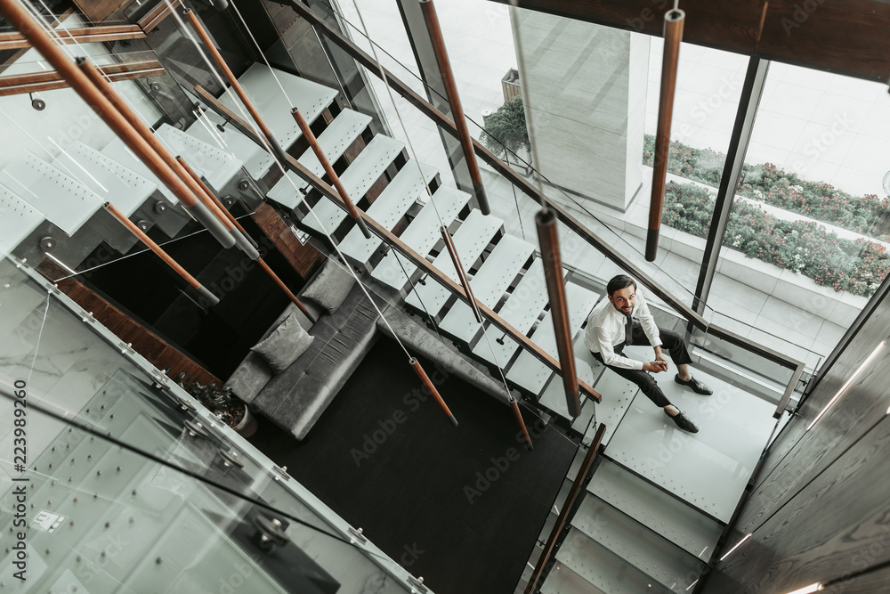 Top view full length portrait of beaming bearded businessman resting on stairs after hard job inside. He looking at camera while locating near wide window