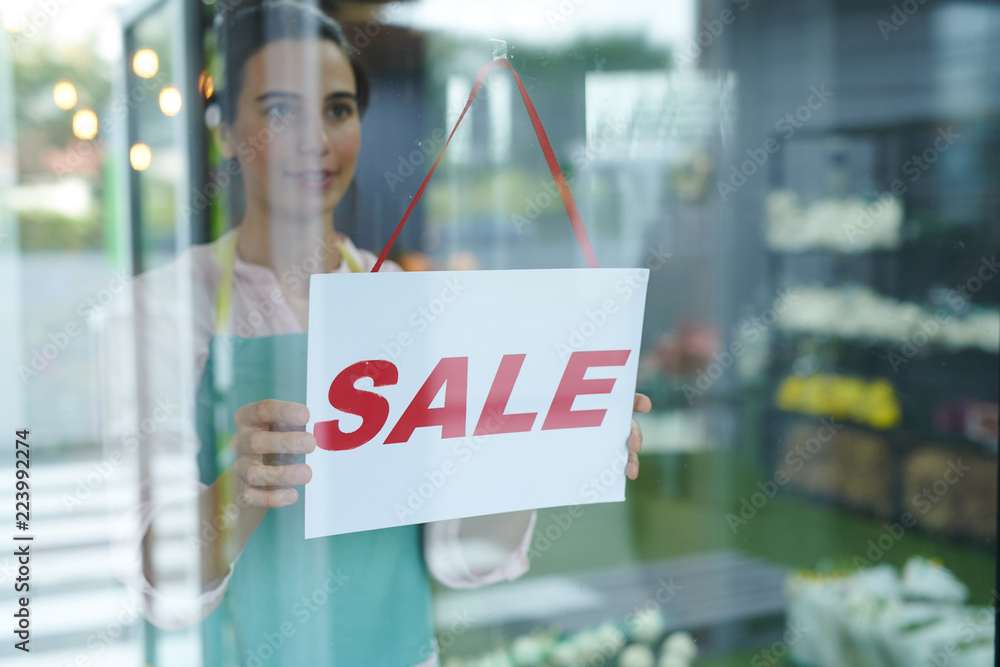 Fototapeta premium Waist up portrait of cheerful female shopkeeper hanging red SALE sign on glass door or window