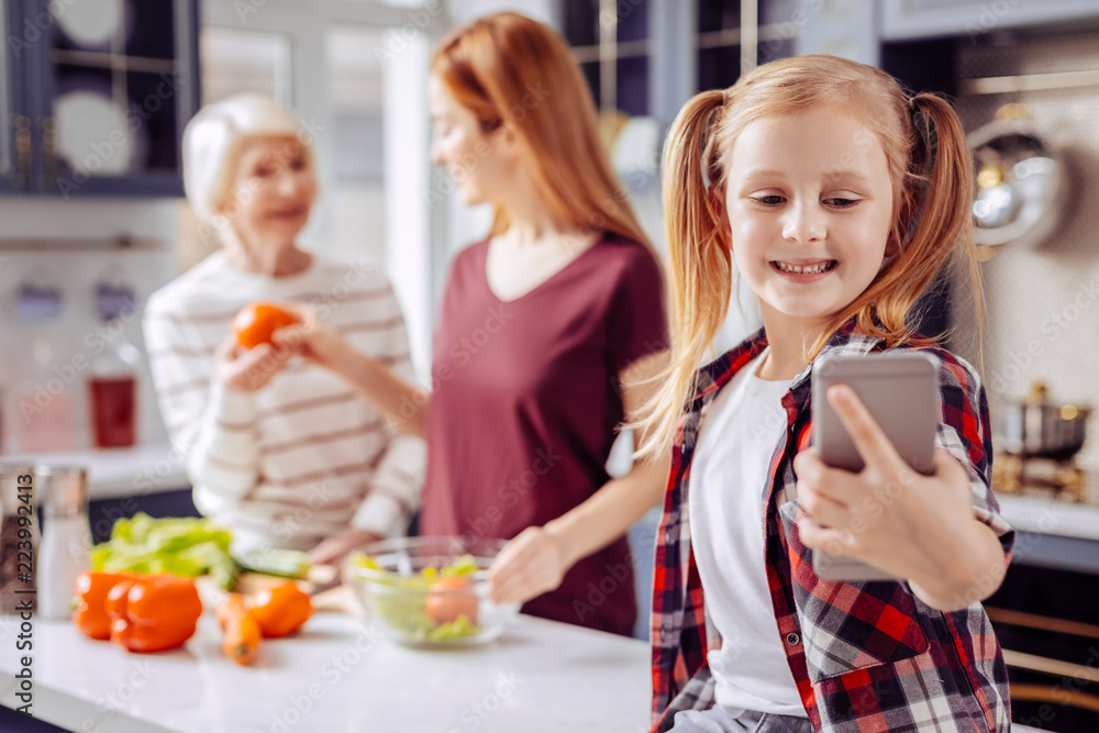 Selfie with family. Cheerful funny little girl holding a modern smartphone and taking selfies with her mother and grandmother in the background