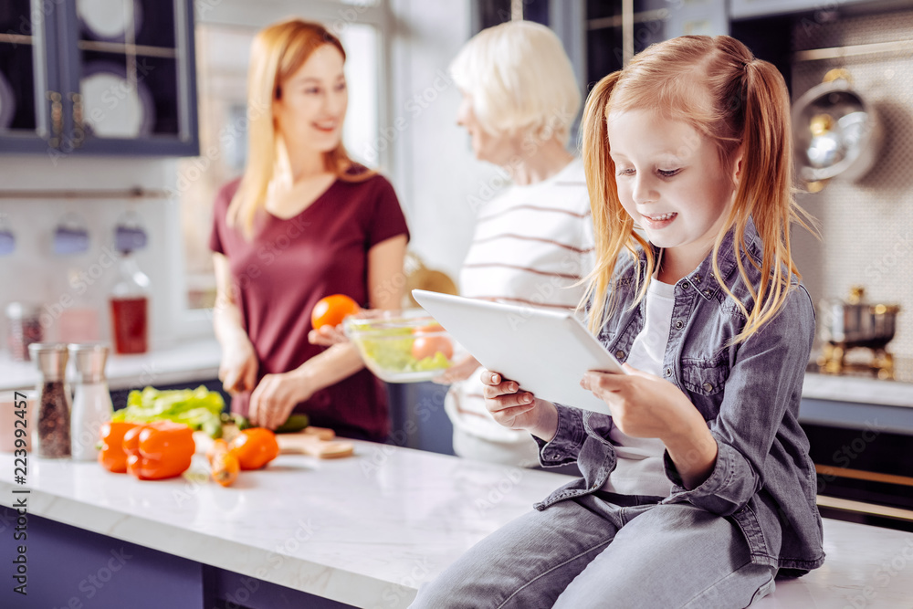 Child with tablet. Cheerful relaxed little girl looking at the screen of a modern tablet while her granny and mother cooking meals in the kitchen Child with tablet. Cheerful relaxed little girl looking at the screen of a modern tablet while her granny and mother cooking meals in the kitchen