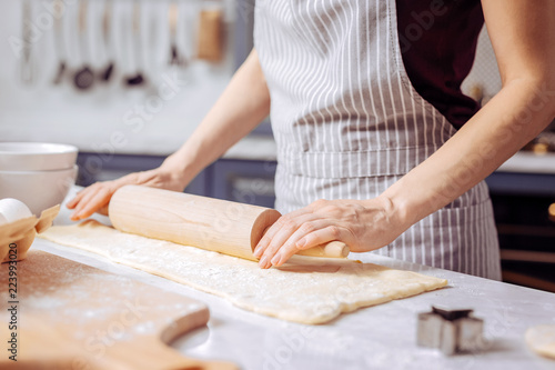 Wooden rolling pin. Careful young woman putting her hands on the wooden rolling pin and using it for working with dough in her kitchen