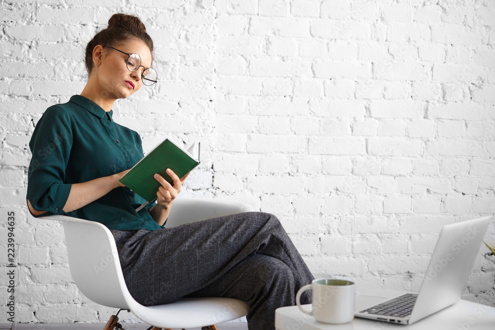 Obraz premium People, job, technology and modern lifestyle concept. Portrait of concentrated serious businesswoman in stylish eyewear working remotely at cafe, writing in diary, sitting with laptop and cup