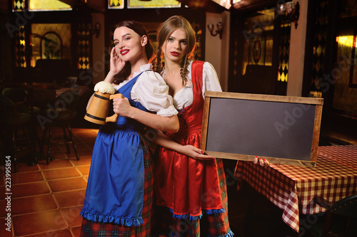 two young attractive girls in traditional Bavarian clothes with chalk Board and a glass of beer in their hands on the background of the pub during the Oktoberfest celebration