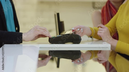 Cheerful customer is using a credit card to pay for purchased clothes while standing at cashier's desk. Shop assistant is accepting transaction and giving paper bags. Man is standing with his wife and