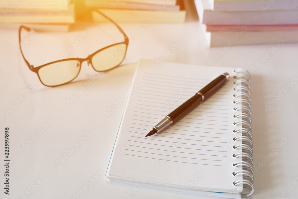 Close up of notebook with ink-pen and eyeglasses put on white table with blur focus of books as a background. Educational concept. Close up of notebook with ink-pen and eyeglasses put on white table with blur focus of books as a background. Educational concept.