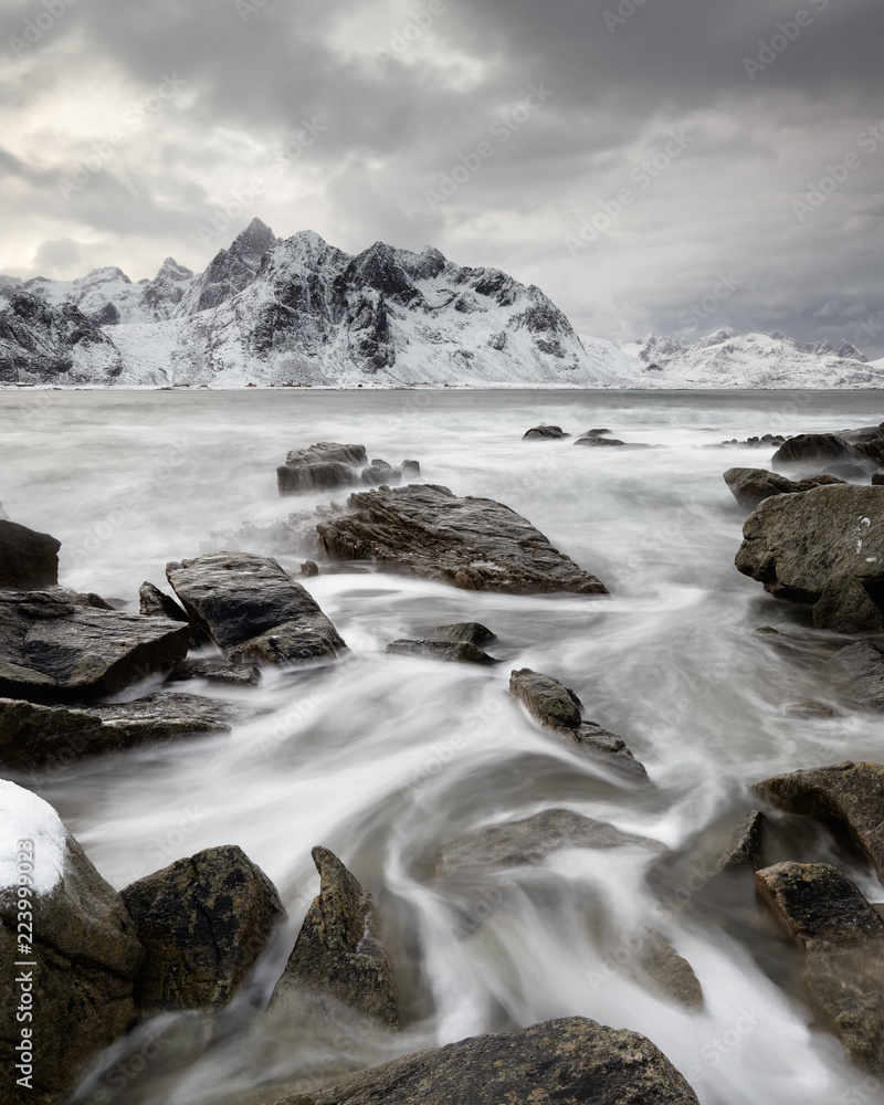 Coastal landscape in winter with water movement between big stones, in the background a mountain range with snow, high contrast sky - Location: Norway, Lofoten