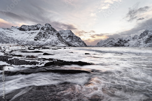Coastal landscape in winter with water movement over large stones, in the background a mountain range with snow, contrasting colorful sky at sunrise - Location: Norway, Lofoten