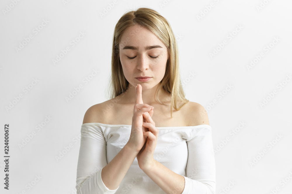 Attractive young female employee meditating in white office, keeping eyes closed and putting hands together in gesture, trying to find balance within herself, practicing breathing exercises Attractive young female employee meditating in white office, keeping eyes closed and putting hands together in gesture, trying to find balance within herself, practicing breathing exercises