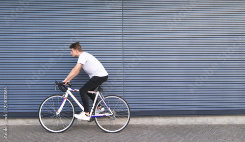 Young man in a white T-shirt rides on a white highway on the background of the wall. Portrait of a student riding a bicycle on the background of the wall. Copyspace