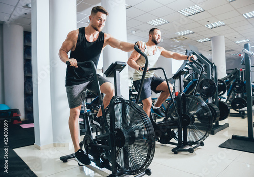 Young men with muscular body using air bike for cardio workout at cross training gym.