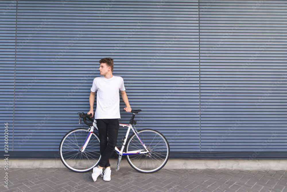 Young student with a white bike stands against the wall and looks to the side. Sports concept. Road bike and young man. Copyspace Young student with a white bike stands against the wall and looks to the side. Sports concept. Road bike and young man. Copyspace