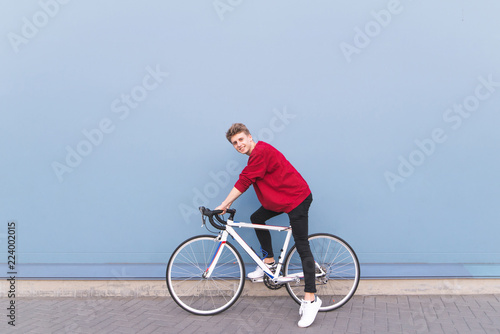 Happy young man in a red sweatshirt sitting on a white bike against a blue wall background. Stylish smiling cyclist on a blue background. Copyspace