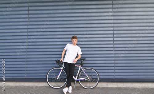 Stylish young man stands with a white highway bike on the background of the wall and looks at the camera. Portrait of a guy with a bicycle on the background of the wall. Copyspace