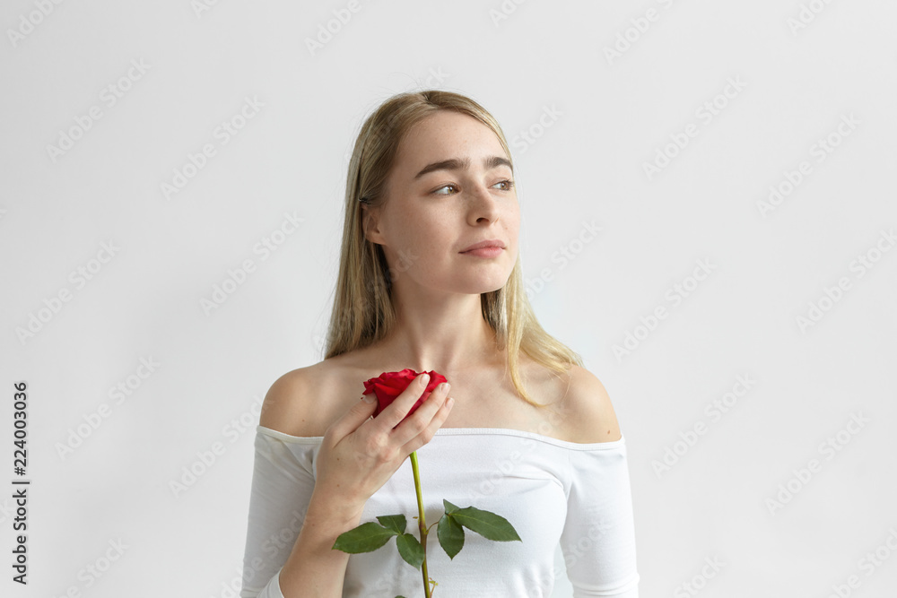 Romantic young Caucasian female wearing open shoulders long sleeved dress holding one red rose from guy on first date, looking sideways with dreamy mysterious smile. Love, passion and romance Romantic young Caucasian female wearing open shoulders long sleeved dress holding one red rose from guy on first date, looking sideways with dreamy mysterious smile. Love, passion and romance