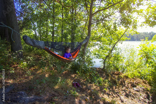 Camping in a hammock. Little girl is resting in a camouflage hammock in a forest near lake at warm autumn sun. Travel and adventure