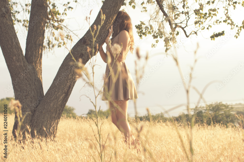girl on nature, young woman walking in summer field at sunset hiding behind a tree