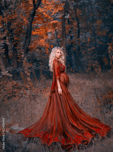 young pregnant woman with blond curly hair in long light flapping red scarlet dress, standing in the forest, holding, hugging her tummy. like a fairy magnificent queen. art autumn dark halloween photo