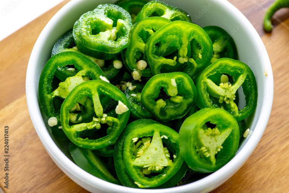 Slicing Jalapeño peppers on a wooden cutting board with a stainless steel knife and placing them in a small white bowl at the end of the board. On a kitchen table. Slicing Jalapeño peppers on a wooden cutting board with a stainless steel knife and placing them in a small white bowl at the end of the board. On a kitchen table.