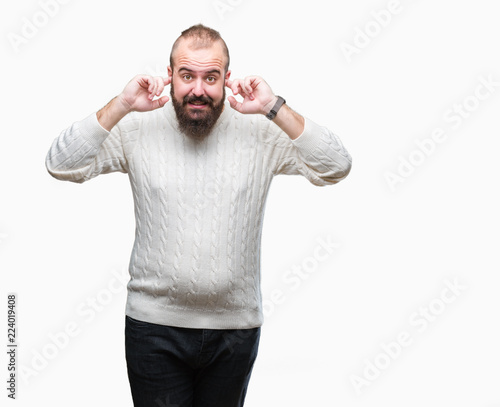 Young caucasian hipster man wearing winter sweater over isolated background covering ears with fingers with annoyed expression for the noise of loud music. Deaf concept.
