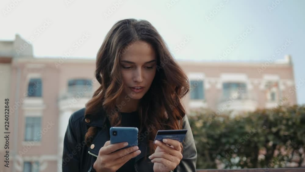 Student on street with credit card and phone Brunette woman sitting on the bench and makes online payments through the Internet from bank card on smartphone Student on street with credit card and phone Brunette woman sitting on the bench and makes online payments through the Internet from bank card on smartphone