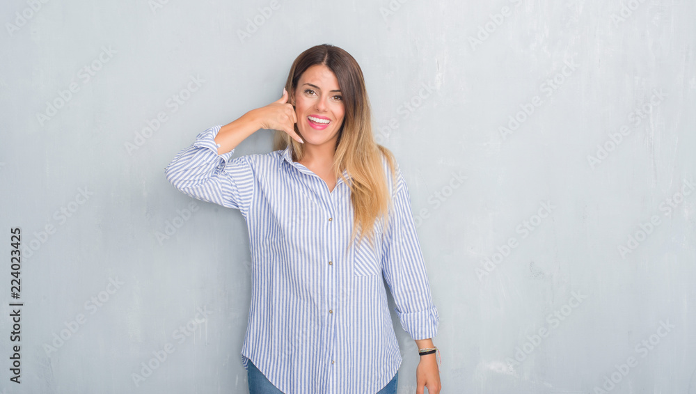 Young adult woman over grey grunge wall wearing fashion business outfit smiling doing phone gesture with hand and fingers like talking on the telephone. Communicating concepts. Young adult woman over grey grunge wall wearing fashion business outfit smiling doing phone gesture with hand and fingers like talking on the telephone. Communicating concepts.