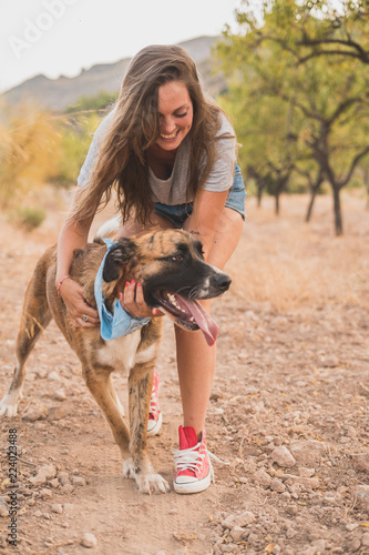 Happy young woman hugging and having fun with her dog with a blue handkerchief around his neck outdoors during sunset. Best friend.