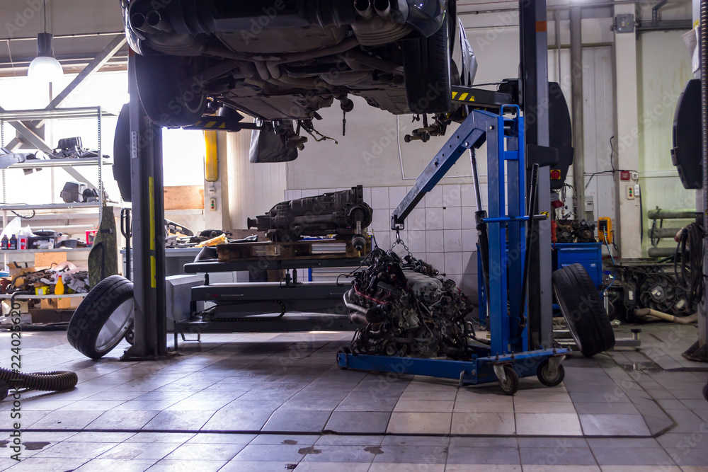 A sports car raised on a lift for repair and under it a detached engine suspended on a blue crane and a gear box on a lifting table in a vehicle repair shop with wheels lying on the sides A sports car raised on a lift for repair and under it a detached engine suspended on a blue crane and a gear box on a lifting table in a vehicle repair shop with wheels lying on the sides