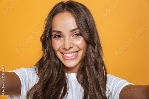 Portrait of happy woman 20s with long hair laughing while taking selfie photo, isolated over yellow background photo