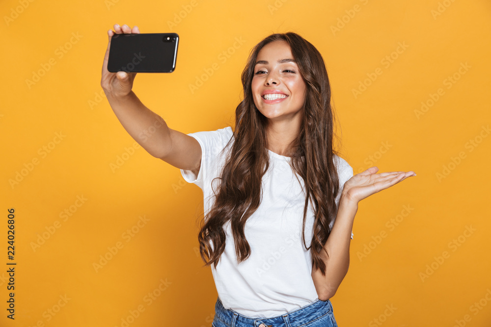 Portrait of positive woman 20s with long hair laughing while taking selfie photo on smartphone, isolated over yellow background Portrait of positive woman 20s with long hair laughing while taking selfie photo on smartphone, isolated over yellow background