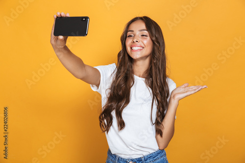 Portrait of positive woman 20s with long hair laughing while taking selfie photo on smartphone, isolated over yellow background