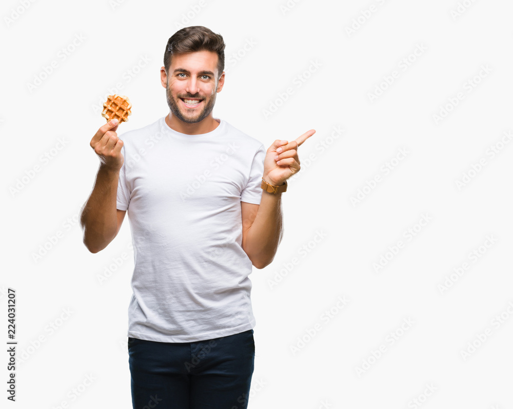 Young handsome man eating a sweet waffle over isolated background very happy pointing with hand and finger to the side Young handsome man eating a sweet waffle over isolated background very happy pointing with hand and finger to the side