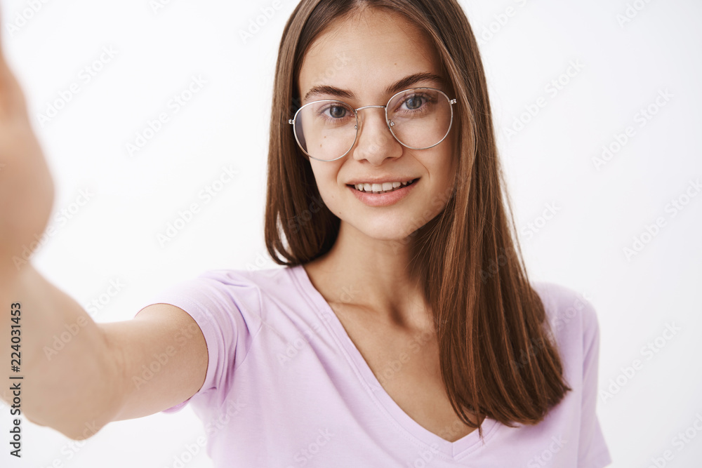 Studio shot of smart attractive and confident adult female entrepreneur in fancy glasses pulling hand forward and smiling at camera while taking selfie or recording video message with new smartphone Studio shot of smart attractive and confident adult female entrepreneur in fancy glasses pulling hand forward and smiling at camera while taking selfie or recording video message with new smartphone