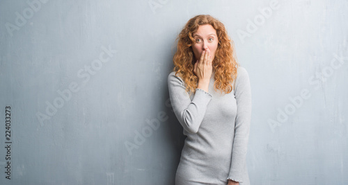 Foto Young redhead woman over grey grunge wall cover mouth with hand shocked with sha