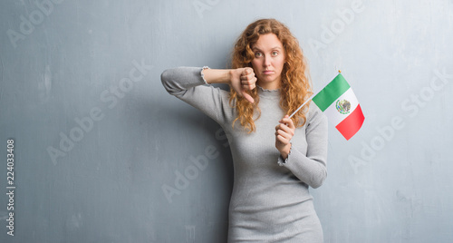 Young redhead woman over grey grunge wall holding flag of Mexico with angry face, negative sign showing dislike with thumbs down, rejection concept