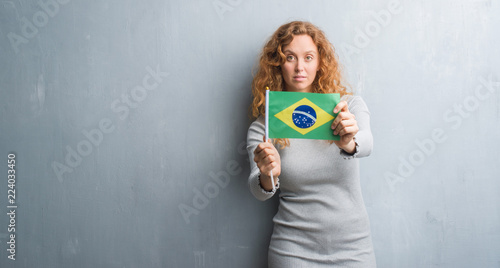 Young redhead woman over grey grunge wall holding flag of Brazil with a confident expression on smart face thinking serious