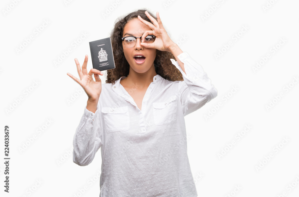 Young hispanic woman holding passport of Canada with happy face smiling doing ok sign with hand on eye looking through fingers Young hispanic woman holding passport of Canada with happy face smiling doing ok sign with hand on eye looking through fingers