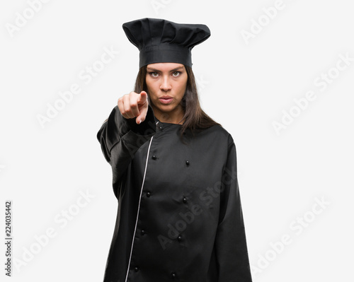 Young hispanic cook woman wearing chef uniform pointing with finger to the camera and to you, hand sign, positive and confident gesture from the front